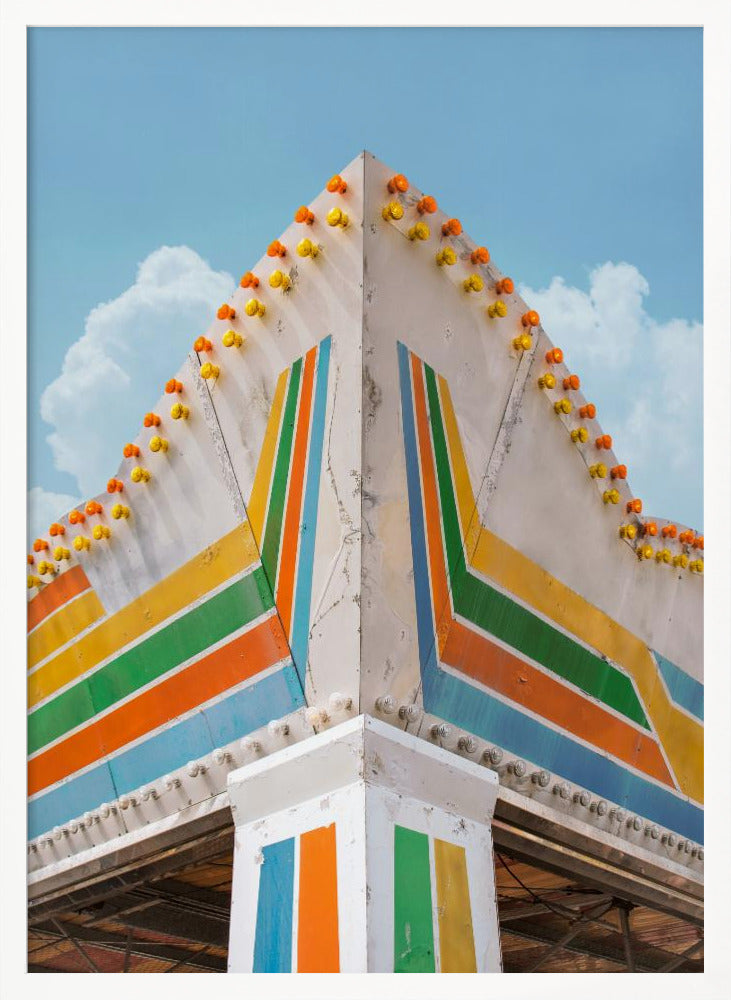 A low-angle photograph of the corner of a retro-style building against a bright blue sky with white clouds. The white facade is painted with bold, colorful stripes in orange, yellow, green, and blue, and the angled roofline is decorated with rows of unlit yellow and orange carnival lights. Print
