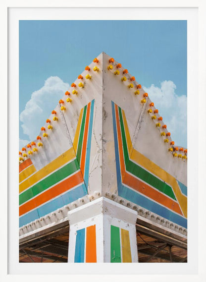 A low-angle photograph of the corner of a retro-style building against a bright blue sky with white clouds. The white facade is painted with bold, colorful stripes in orange, yellow, green, and blue, and the angled roofline is decorated with rows of unlit yellow and orange carnival lights. Print