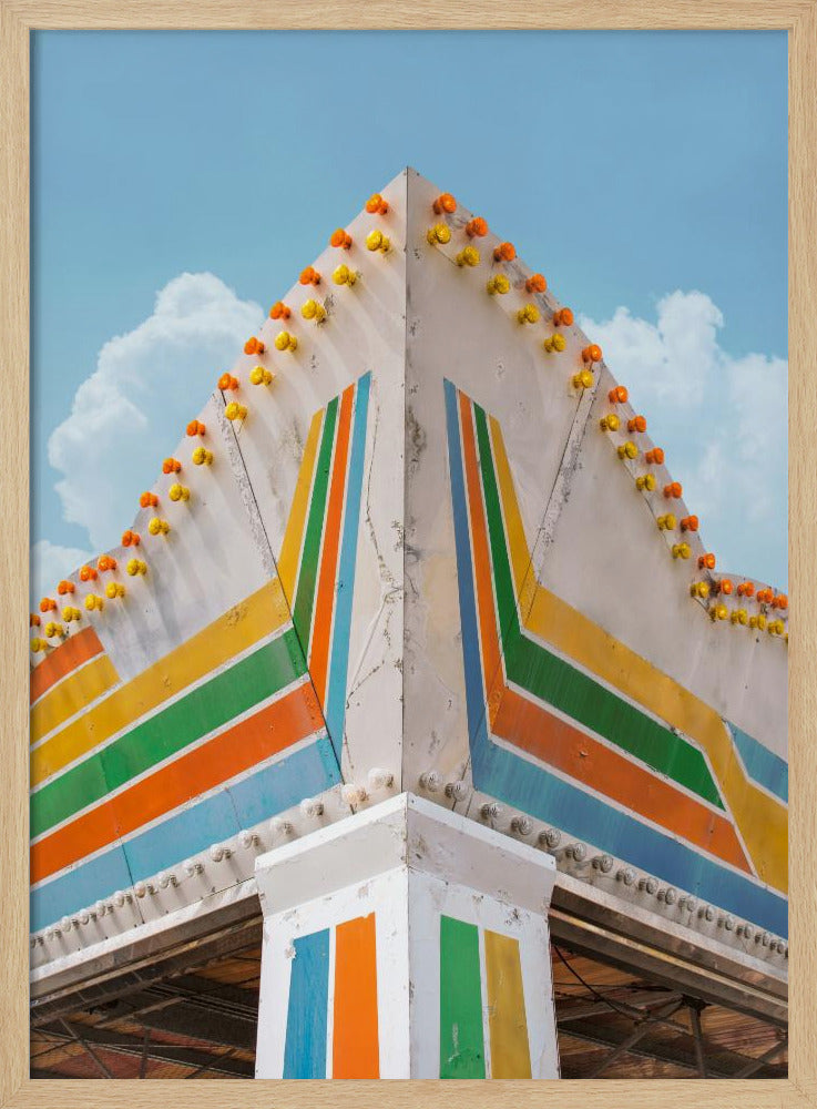 A low-angle photograph of the corner of a retro-style building against a bright blue sky with white clouds. The white facade is painted with bold, colorful stripes in orange, yellow, green, and blue, and the angled roofline is decorated with rows of unlit yellow and orange carnival lights. Print