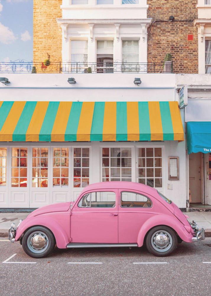 A vibrant, eye-level photograph of a vintage pink Volkswagen Beetle parked on a city street in front of a charming white storefront with a bold yellow and teal striped awning. Wall Art