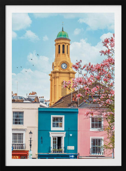 A vibrant city street scene on a sunny day, featuring a row of colorful houses in teal, pink, and off-white. A striking yellow clock tower with a green dome stands tall against a bright blue sky, with a blooming pink magnolia tree in the foreground. Print