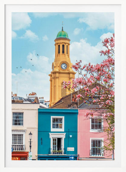 A vibrant city street scene on a sunny day, featuring a row of colorful houses in teal, pink, and off-white. A striking yellow clock tower with a green dome stands tall against a bright blue sky, with a blooming pink magnolia tree in the foreground. Print