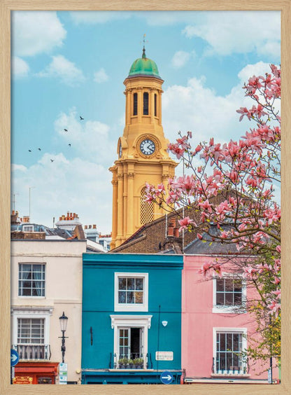 A vibrant city street scene on a sunny day, featuring a row of colorful houses in teal, pink, and off-white. A striking yellow clock tower with a green dome stands tall against a bright blue sky, with a blooming pink magnolia tree in the foreground. Print