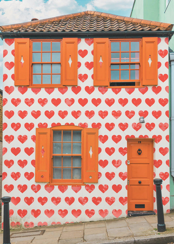 The front facade of a whimsical two-story house with white walls covered in a pattern of red hearts. The house has bright orange shutters on its windows and a matching orange front door, creating a vibrant and playful look against the heart-patterned background. Artwork