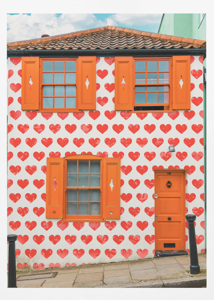 The front facade of a whimsical two-story house with white walls covered in a pattern of red hearts. The house has bright orange shutters on its windows and a matching orange front door, creating a vibrant and playful look against the heart-patterned background. Artwork