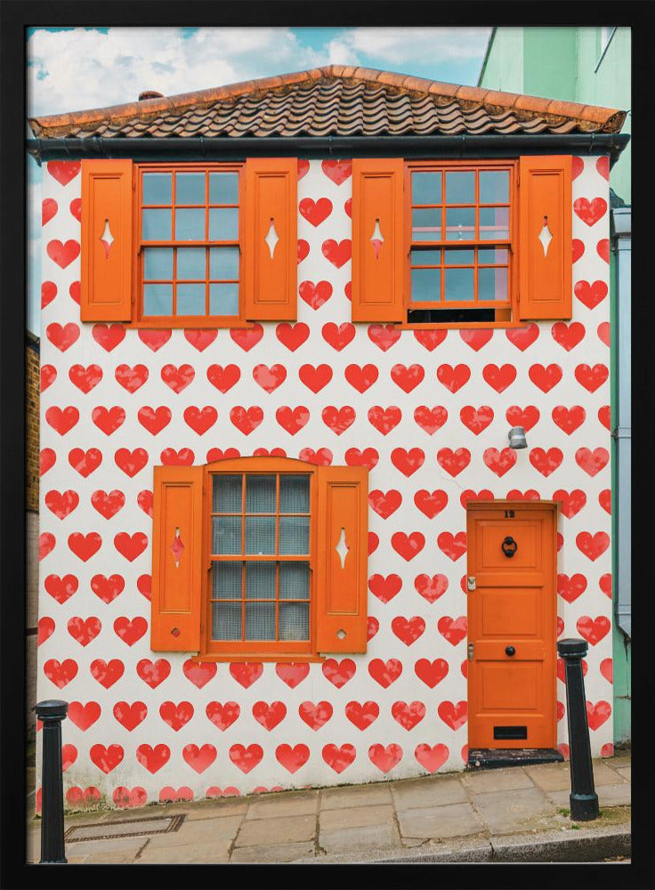 The front facade of a whimsical two-story house with white walls covered in a pattern of red hearts. The house has bright orange shutters on its windows and a matching orange front door, creating a vibrant and playful look against the heart-patterned background. Artwork