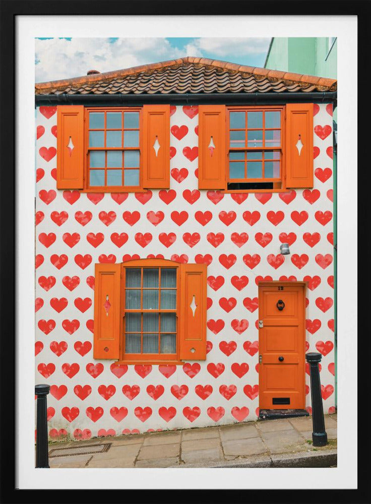 The front facade of a whimsical two-story house with white walls covered in a pattern of red hearts. The house has bright orange shutters on its windows and a matching orange front door, creating a vibrant and playful look against the heart-patterned background. Artwork