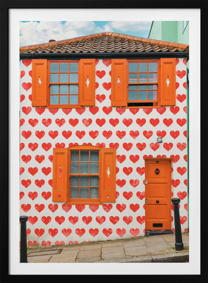 The front facade of a whimsical two-story house with white walls covered in a pattern of red hearts. The house has bright orange shutters on its windows and a matching orange front door, creating a vibrant and playful look against the heart-patterned background. Artwork
