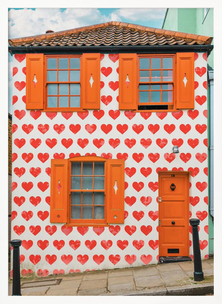 The front facade of a whimsical two-story house with white walls covered in a pattern of red hearts. The house has bright orange shutters on its windows and a matching orange front door, creating a vibrant and playful look against the heart-patterned background. Artwork