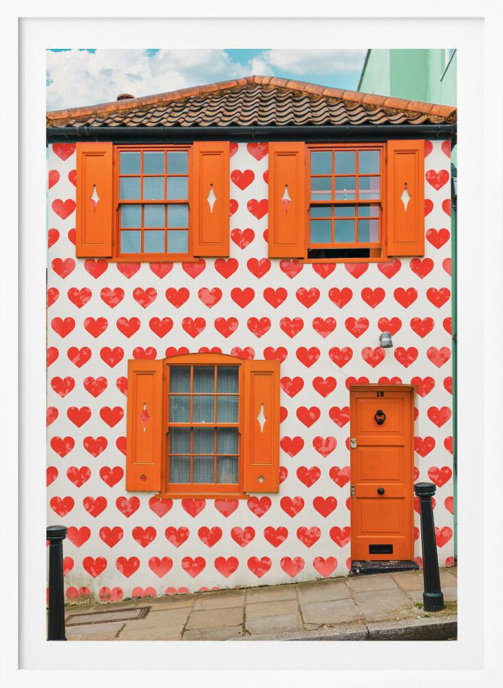 The front facade of a whimsical two-story house with white walls covered in a pattern of red hearts. The house has bright orange shutters on its windows and a matching orange front door, creating a vibrant and playful look against the heart-patterned background. Artwork
