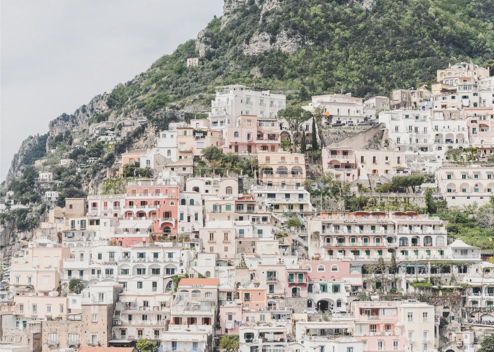 Photograph of the iconic cliffside village of Positano on the Amalfi Coast, showcasing a dense cascade of white, pink, and peach buildings down a lush green mountain under an overcast sky. The image is presented within a silver picture frame. Artwork