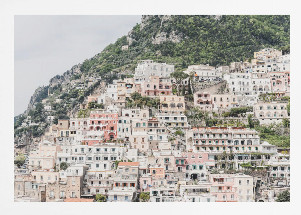 Photograph of the iconic cliffside village of Positano on the Amalfi Coast, showcasing a dense cascade of white, pink, and peach buildings down a lush green mountain under an overcast sky. The image is presented within a silver picture frame. Artwork