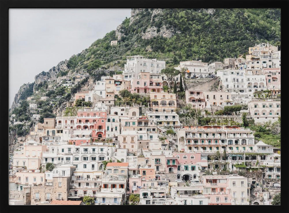 Photograph of the iconic cliffside village of Positano on the Amalfi Coast, showcasing a dense cascade of white, pink, and peach buildings down a lush green mountain under an overcast sky. The image is presented within a silver picture frame. Artwork