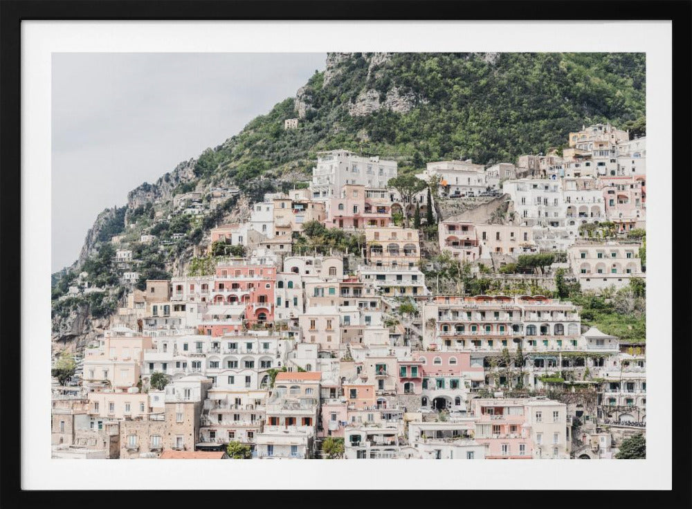 Photograph of the iconic cliffside village of Positano on the Amalfi Coast, showcasing a dense cascade of white, pink, and peach buildings down a lush green mountain under an overcast sky. The image is presented within a silver picture frame. Artwork