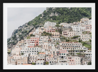 Photograph of the iconic cliffside village of Positano on the Amalfi Coast, showcasing a dense cascade of white, pink, and peach buildings down a lush green mountain under an overcast sky. The image is presented within a silver picture frame. Artwork
