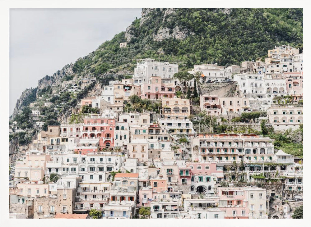 Photograph of the iconic cliffside village of Positano on the Amalfi Coast, showcasing a dense cascade of white, pink, and peach buildings down a lush green mountain under an overcast sky. The image is presented within a silver picture frame. Artwork