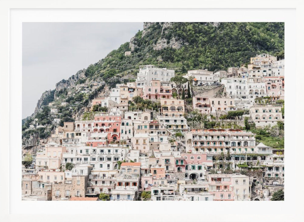 Photograph of the iconic cliffside village of Positano on the Amalfi Coast, showcasing a dense cascade of white, pink, and peach buildings down a lush green mountain under an overcast sky. The image is presented within a silver picture frame. Artwork