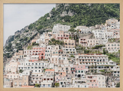 Photograph of the iconic cliffside village of Positano on the Amalfi Coast, showcasing a dense cascade of white, pink, and peach buildings down a lush green mountain under an overcast sky. The image is presented within a silver picture frame. Artwork