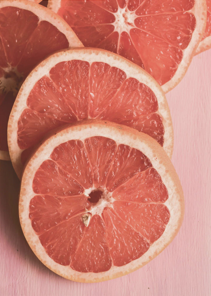 A close-up photograph of several overlapping slices of vibrant pink grapefruit arranged on a pale pink surface, enclosed within a simple black frame. Print