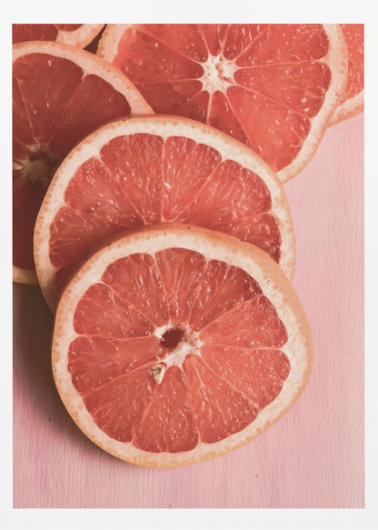A close-up photograph of several overlapping slices of vibrant pink grapefruit arranged on a pale pink surface, enclosed within a simple black frame. Print