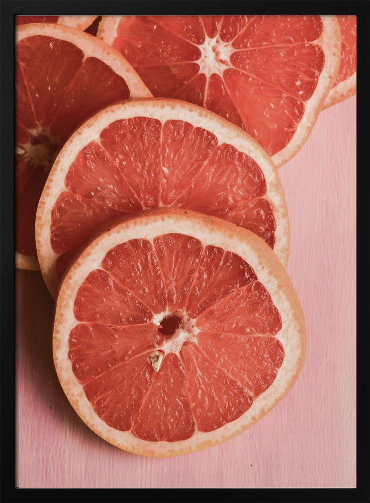 A close-up photograph of several overlapping slices of vibrant pink grapefruit arranged on a pale pink surface, enclosed within a simple black frame. Print