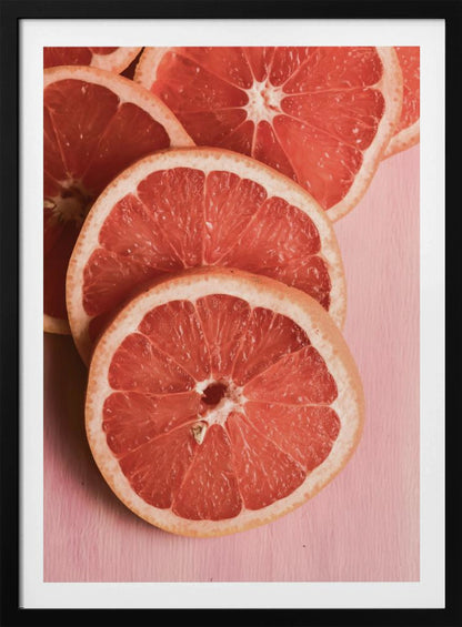 A close-up photograph of several overlapping slices of vibrant pink grapefruit arranged on a pale pink surface, enclosed within a simple black frame. Print