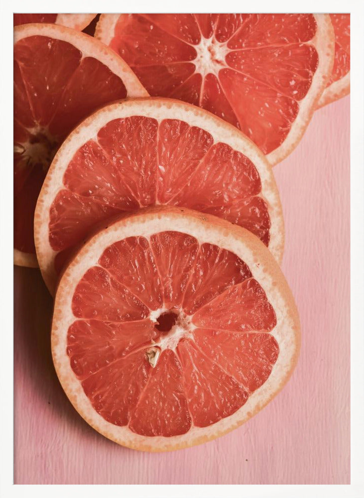 A close-up photograph of several overlapping slices of vibrant pink grapefruit arranged on a pale pink surface, enclosed within a simple black frame. Print