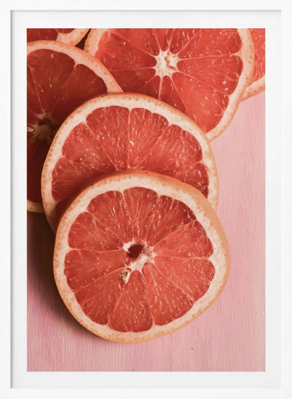 A close-up photograph of several overlapping slices of vibrant pink grapefruit arranged on a pale pink surface, enclosed within a simple black frame. Print