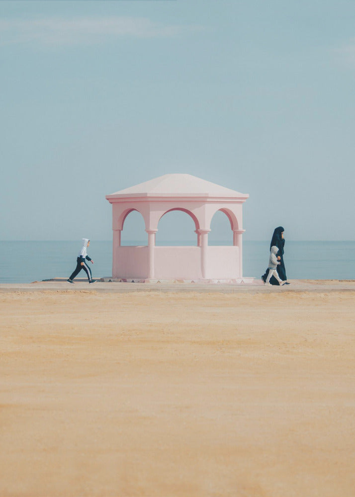 A minimalist photograph of a coastal scene with a pastel color palette. A pink arched gazebo sits on a promenade with the blue sea and sky in the background. A large expanse of beige sand is in the foreground. Two figures in hijabs walk in opposite directions on either side of the gazebo, one with a small child. Decor