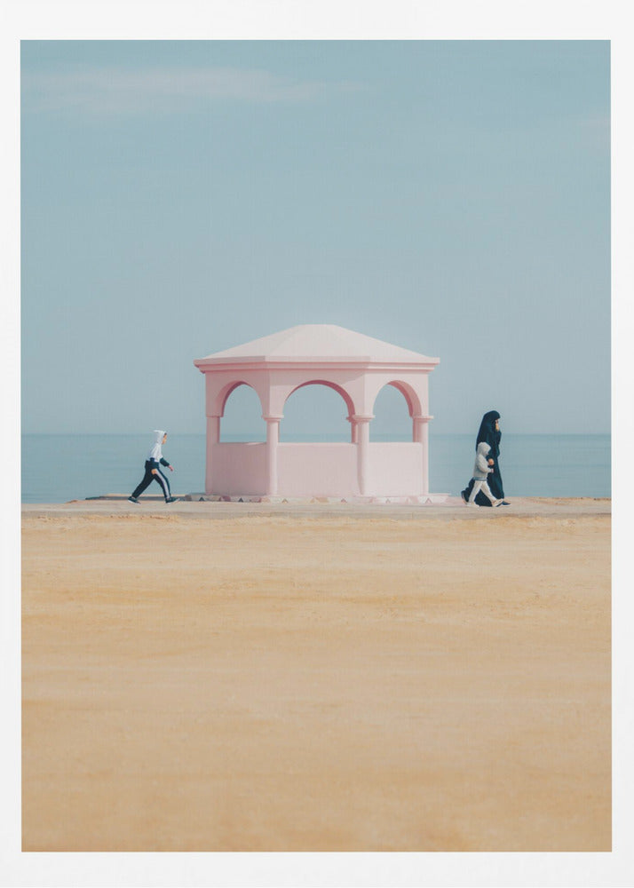 A minimalist photograph of a coastal scene with a pastel color palette. A pink arched gazebo sits on a promenade with the blue sea and sky in the background. A large expanse of beige sand is in the foreground. Two figures in hijabs walk in opposite directions on either side of the gazebo, one with a small child. Decor