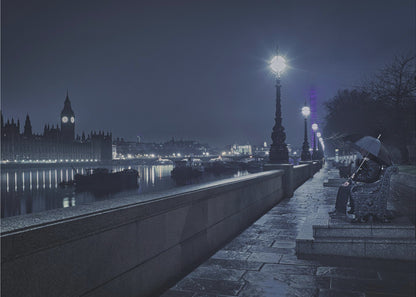 A moody, blue-toned night photograph of London. A solitary figure sits on a bench under an umbrella on a wet stone walkway along the River Thames, illuminated by vintage streetlights. Across the river, the iconic Houses of Parliament and Big Ben are lit up against the dark sky, their lights reflecting in the water. Poster