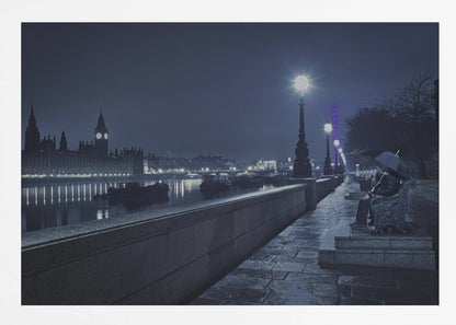 A moody, blue-toned night photograph of London. A solitary figure sits on a bench under an umbrella on a wet stone walkway along the River Thames, illuminated by vintage streetlights. Across the river, the iconic Houses of Parliament and Big Ben are lit up against the dark sky, their lights reflecting in the water. Poster