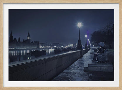 A moody, blue-toned night photograph of London. A solitary figure sits on a bench under an umbrella on a wet stone walkway along the River Thames, illuminated by vintage streetlights. Across the river, the iconic Houses of Parliament and Big Ben are lit up against the dark sky, their lights reflecting in the water. Poster