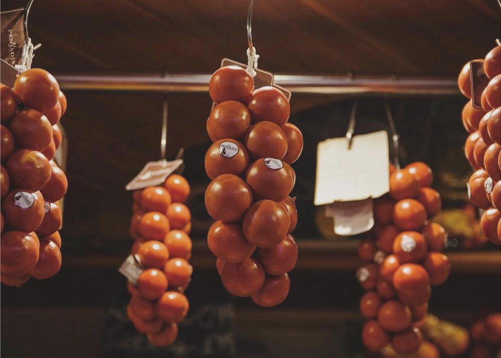A close-up, warm-toned photograph of several bunches of ripe red tomatoes hanging from hooks in a row, likely at a market or in a pantry. The background is dark and out of focus, highlighting the vibrant produce. Poster