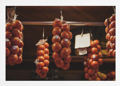 A close-up, warm-toned photograph of several bunches of ripe red tomatoes hanging from hooks in a row, likely at a market or in a pantry. The background is dark and out of focus, highlighting the vibrant produce. Poster