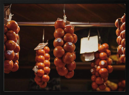 A close-up, warm-toned photograph of several bunches of ripe red tomatoes hanging from hooks in a row, likely at a market or in a pantry. The background is dark and out of focus, highlighting the vibrant produce. Poster