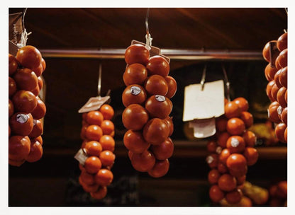 A close-up, warm-toned photograph of several bunches of ripe red tomatoes hanging from hooks in a row, likely at a market or in a pantry. The background is dark and out of focus, highlighting the vibrant produce. Poster