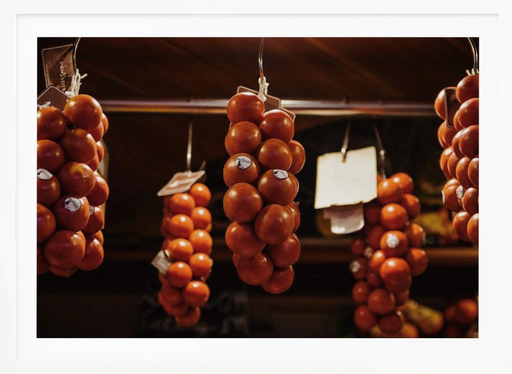 A close-up, warm-toned photograph of several bunches of ripe red tomatoes hanging from hooks in a row, likely at a market or in a pantry. The background is dark and out of focus, highlighting the vibrant produce. Poster