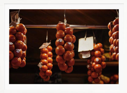 A close-up, warm-toned photograph of several bunches of ripe red tomatoes hanging from hooks in a row, likely at a market or in a pantry. The background is dark and out of focus, highlighting the vibrant produce. Poster