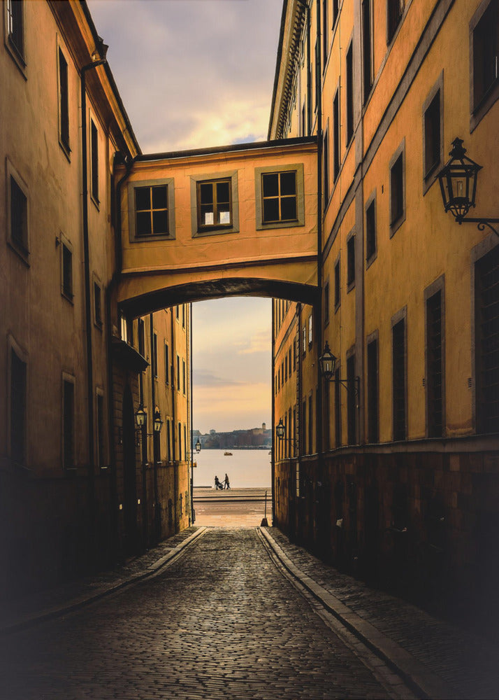 A perspective view down a narrow, shadowy cobblestone alley in an old European city, framed by warm, ochre-colored buildings. An arched walkway connects the buildings overhead, and the alley opens to a view of a tranquil body of water at sunset, with two small figures silhouetted by the shore. Decor