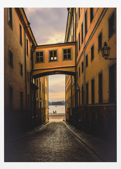 A perspective view down a narrow, shadowy cobblestone alley in an old European city, framed by warm, ochre-colored buildings. An arched walkway connects the buildings overhead, and the alley opens to a view of a tranquil body of water at sunset, with two small figures silhouetted by the shore. Decor