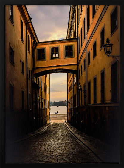 A perspective view down a narrow, shadowy cobblestone alley in an old European city, framed by warm, ochre-colored buildings. An arched walkway connects the buildings overhead, and the alley opens to a view of a tranquil body of water at sunset, with two small figures silhouetted by the shore. Decor