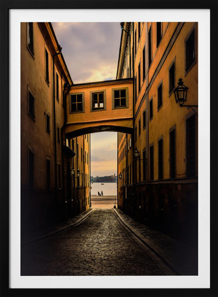A perspective view down a narrow, shadowy cobblestone alley in an old European city, framed by warm, ochre-colored buildings. An arched walkway connects the buildings overhead, and the alley opens to a view of a tranquil body of water at sunset, with two small figures silhouetted by the shore. Decor