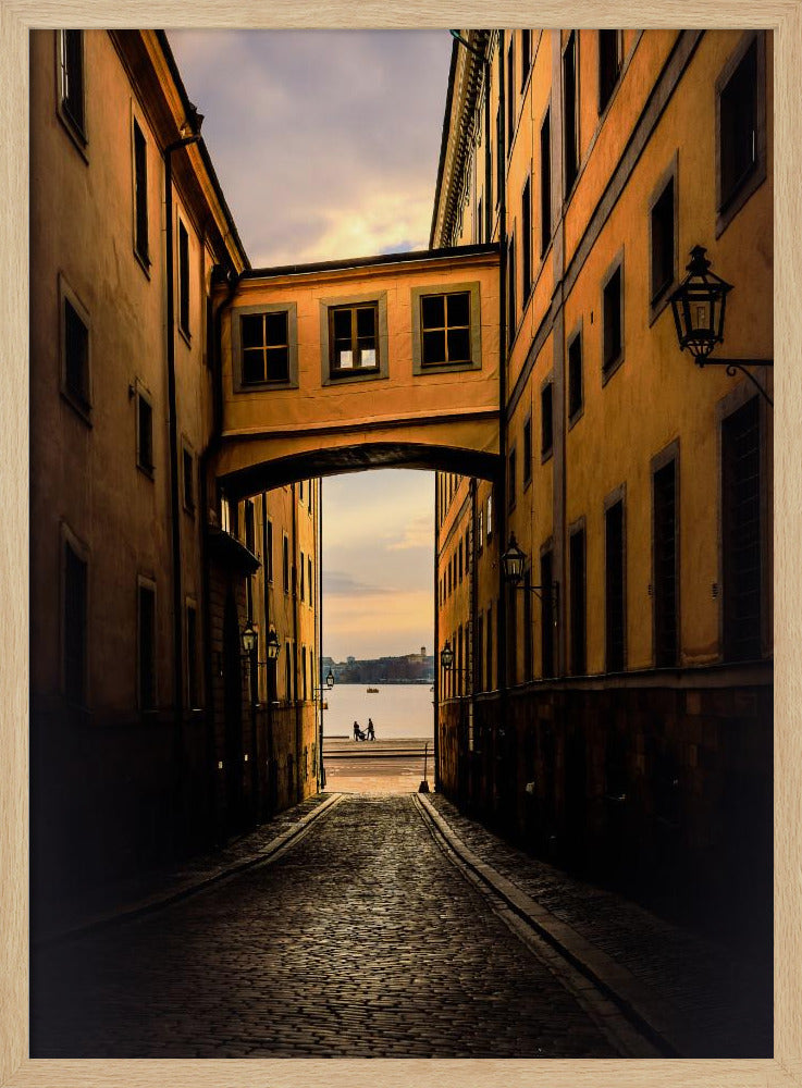 A perspective view down a narrow, shadowy cobblestone alley in an old European city, framed by warm, ochre-colored buildings. An arched walkway connects the buildings overhead, and the alley opens to a view of a tranquil body of water at sunset, with two small figures silhouetted by the shore. Decor
