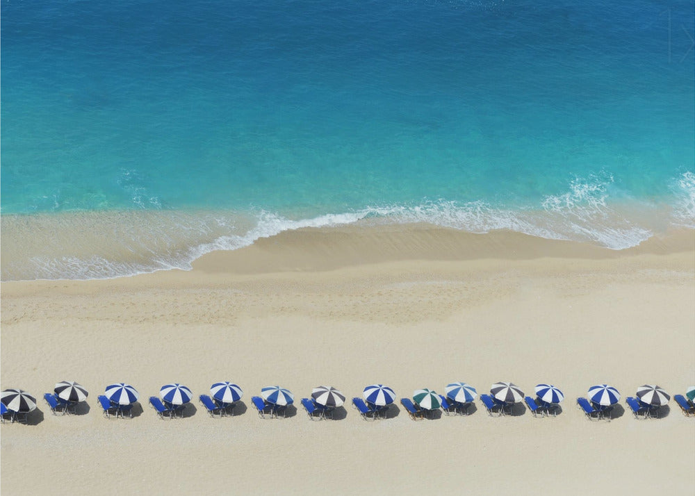 An aerial view of a pristine sandy beach with vibrant turquoise ocean waves gently lapping the shore. A long, neat row of blue and white striped beach umbrellas stands along the sand, creating a serene and orderly coastal scene, all enclosed within a silver frame. Artwork