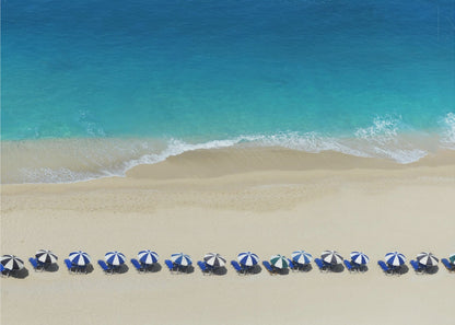 An aerial view of a pristine sandy beach with vibrant turquoise ocean waves gently lapping the shore. A long, neat row of blue and white striped beach umbrellas stands along the sand, creating a serene and orderly coastal scene, all enclosed within a silver frame. Artwork