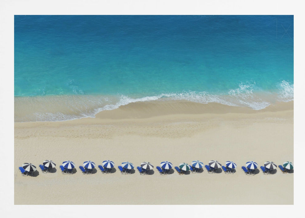 An aerial view of a pristine sandy beach with vibrant turquoise ocean waves gently lapping the shore. A long, neat row of blue and white striped beach umbrellas stands along the sand, creating a serene and orderly coastal scene, all enclosed within a silver frame. Artwork