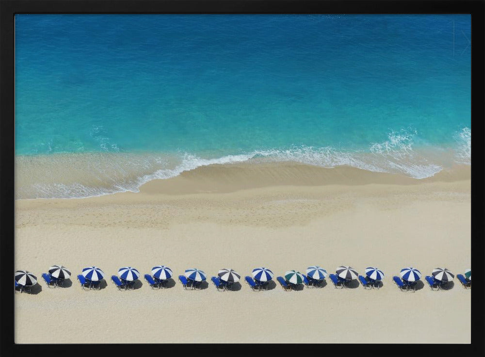 An aerial view of a pristine sandy beach with vibrant turquoise ocean waves gently lapping the shore. A long, neat row of blue and white striped beach umbrellas stands along the sand, creating a serene and orderly coastal scene, all enclosed within a silver frame. Artwork
