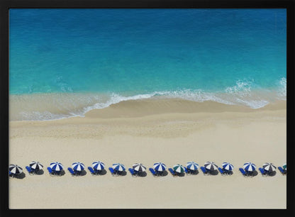 An aerial view of a pristine sandy beach with vibrant turquoise ocean waves gently lapping the shore. A long, neat row of blue and white striped beach umbrellas stands along the sand, creating a serene and orderly coastal scene, all enclosed within a silver frame. Artwork