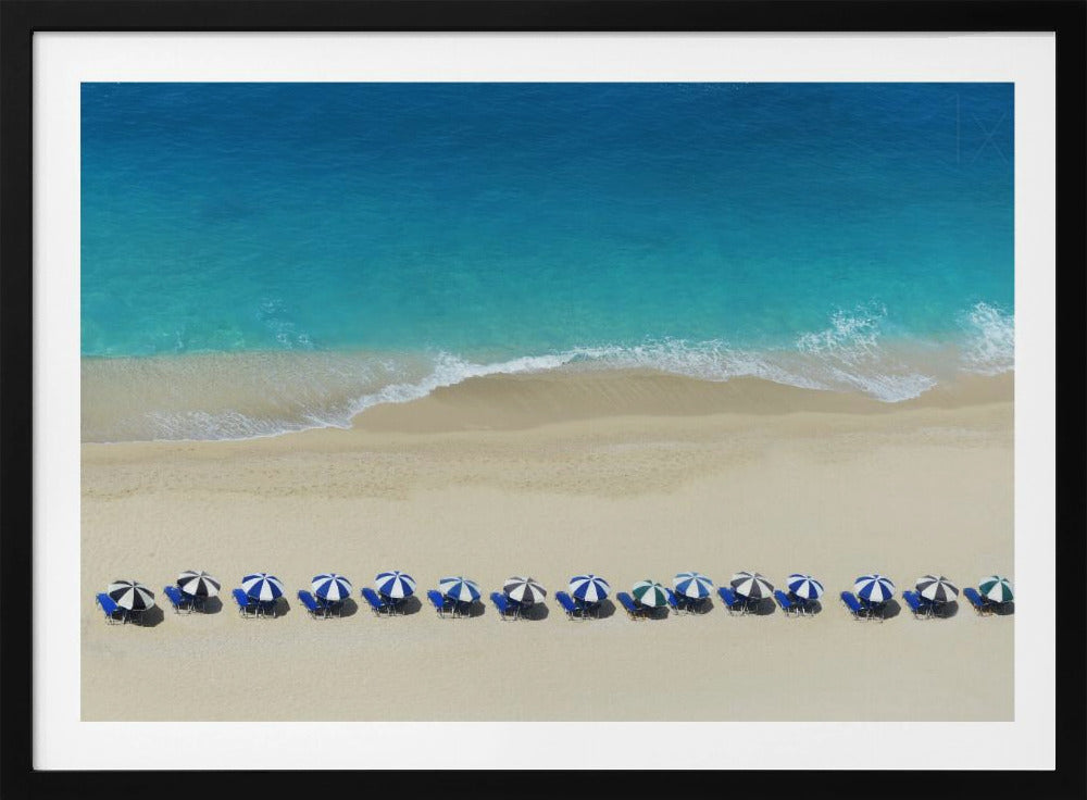 An aerial view of a pristine sandy beach with vibrant turquoise ocean waves gently lapping the shore. A long, neat row of blue and white striped beach umbrellas stands along the sand, creating a serene and orderly coastal scene, all enclosed within a silver frame. Artwork